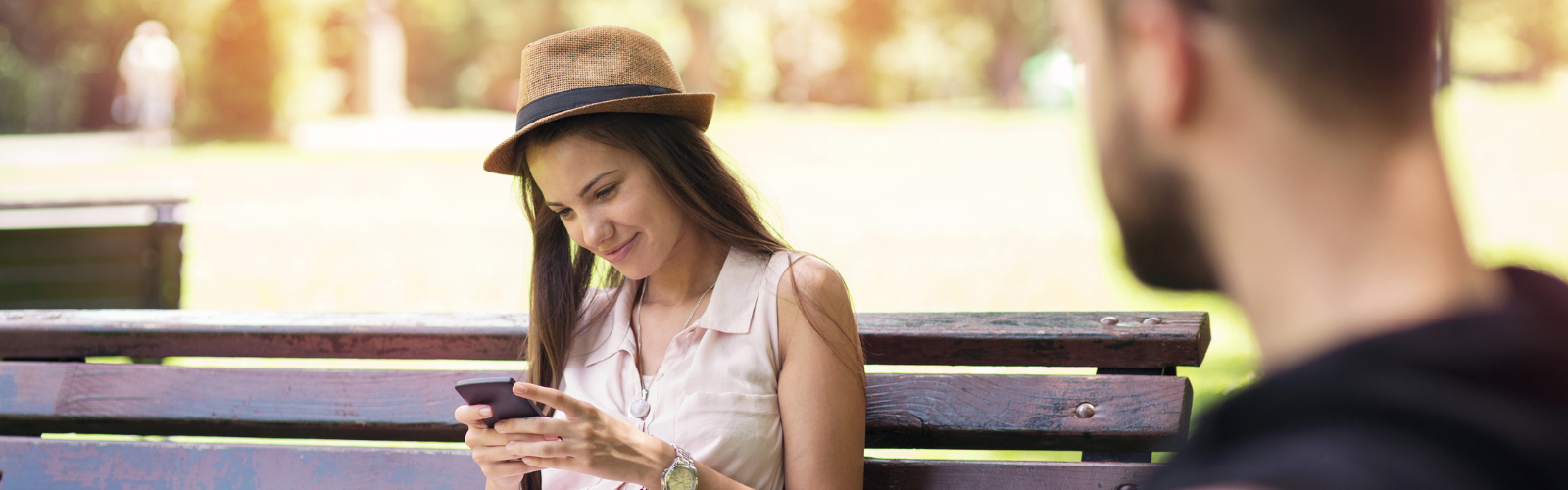 Frau mit Hut sitzt auf einer Parkbank und lächelt beim Blick auf ihr Handy, ein Mann unscharf im Vordergrund.