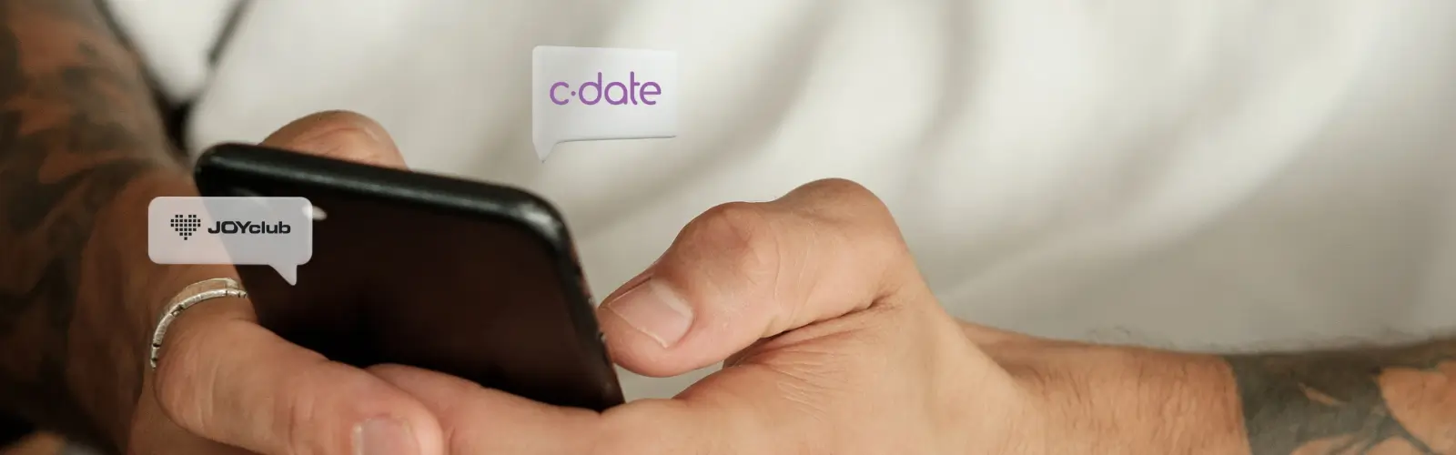 Nahaufnahme eines Mannes mit Tätowierungen, der auf seinem Smartphone scrollt. Über dem Bildschirm sind die Logos von C-Date und JOYclub eingeblendet.