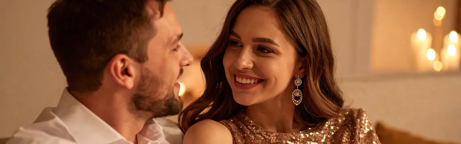 Ein lächelndes Paar in festlicher Kleidung, das sich bei gedämpftem Kerzenlicht liebevoll ansieht. Die Frau trägt ein glitzerndes Paillettenkleid.