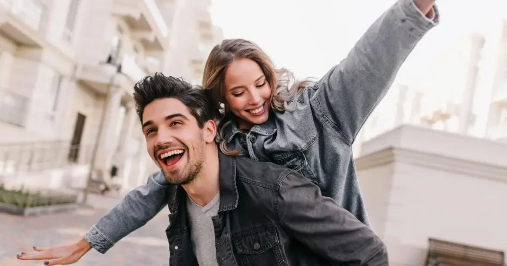 Glückliches junges Paar bei einem fröhlichen Piggyback-Ritt im Freien, beide lächeln in lässigen Denim-Jacken – Prestigemen