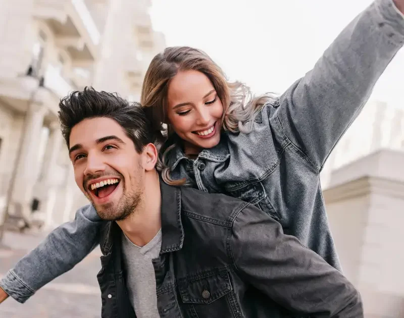 Glückliches junges Paar bei einem fröhlichen Piggyback-Ritt im Freien, beide lächeln in lässigen Denim-Jacken – Prestigemen