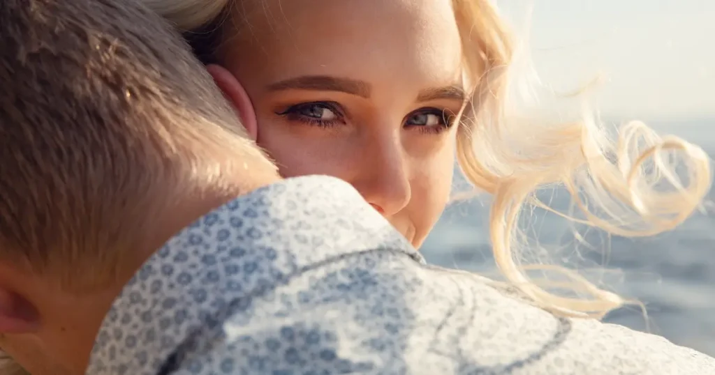 Frau umarmt ihren Partner liebevoll von hinten und blickt zufrieden mit einem kleinen Lächeln über seine Schulter.