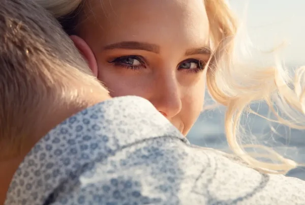 Frau umarmt ihren Partner liebevoll von hinten und blickt zufrieden mit einem kleinen Lächeln über seine Schulter.