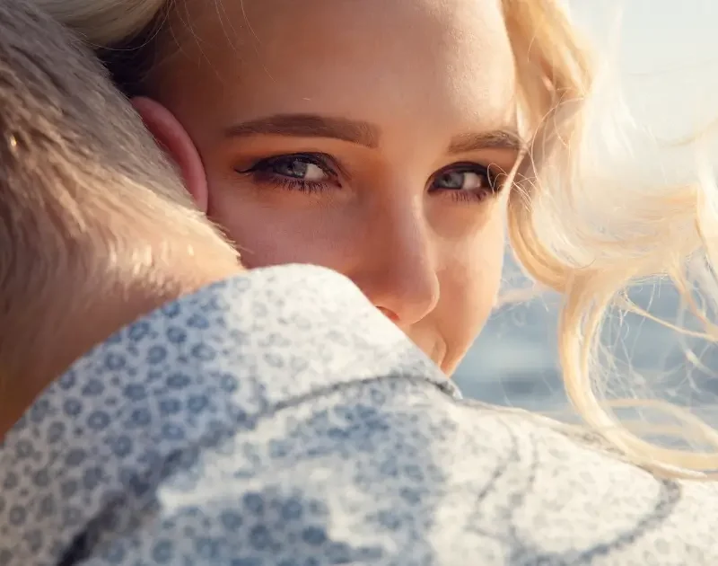 Frau umarmt ihren Partner liebevoll von hinten und blickt zufrieden mit einem kleinen Lächeln über seine Schulter.