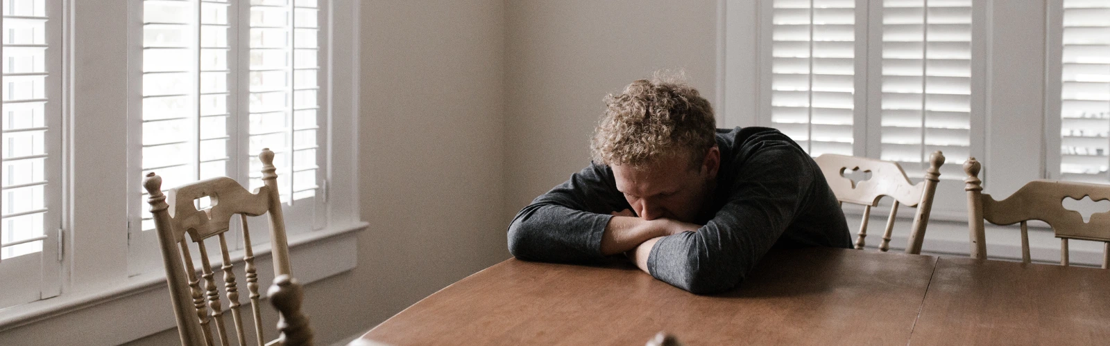 Ein erschöpfter Mann lehnt seinen Kopf in einem hellen Raum auf seine Arme am Esstisch und zeigt Anzeichen von Trauer und Rückzug.