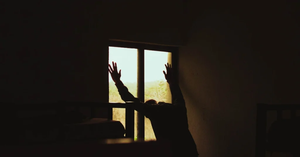 Silhouette eines verzweifelten Mannes, der seine Hände gegen ein Fenster drückt, als Symbol für den Schmerz während der ersten Phase des Liebeskummers.