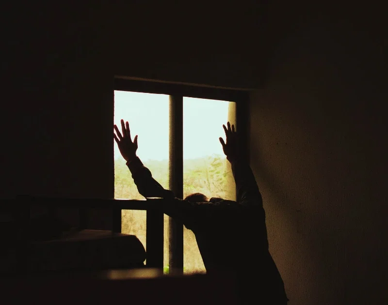 Silhouette eines verzweifelten Mannes, der seine Hände gegen ein Fenster drückt, als Symbol für den Schmerz während der ersten Phase des Liebeskummers.