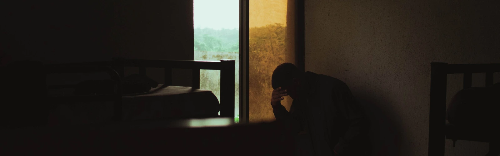 Ein Mann sitzt nachdenklich im Schatten am Fenster und hält sich den Kopf, was die tiefe Reflexion über die eigene Trennungsphase darstellt.
