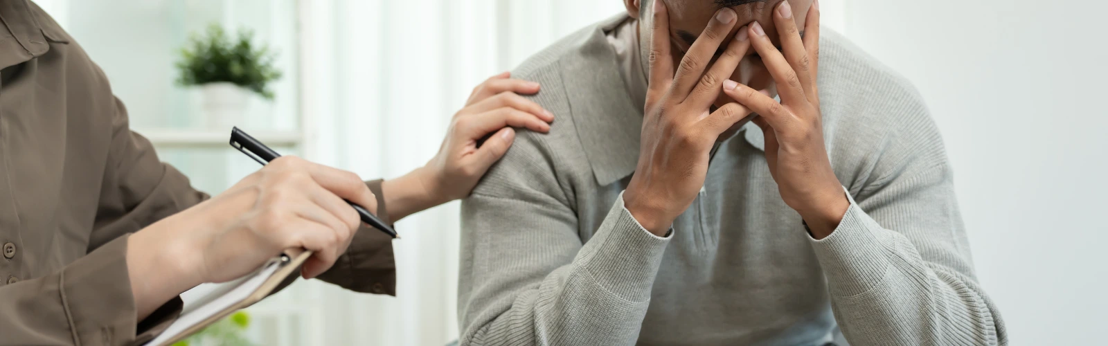 Ein verzweifelter Mann hält sein Gesicht in den Händen, während eine Therapeutin tröstend eine Hand auf seine Schulter legt.