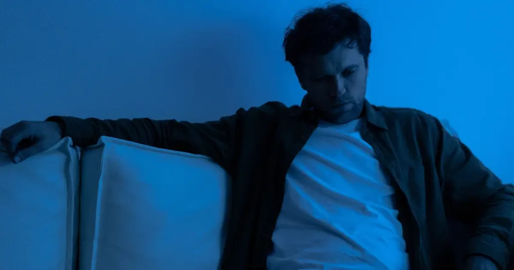 Nachdenklicher Mann sitzt allein auf dem Sofa in blauem Licht, Sinnbild für emotionale Rückfälle.