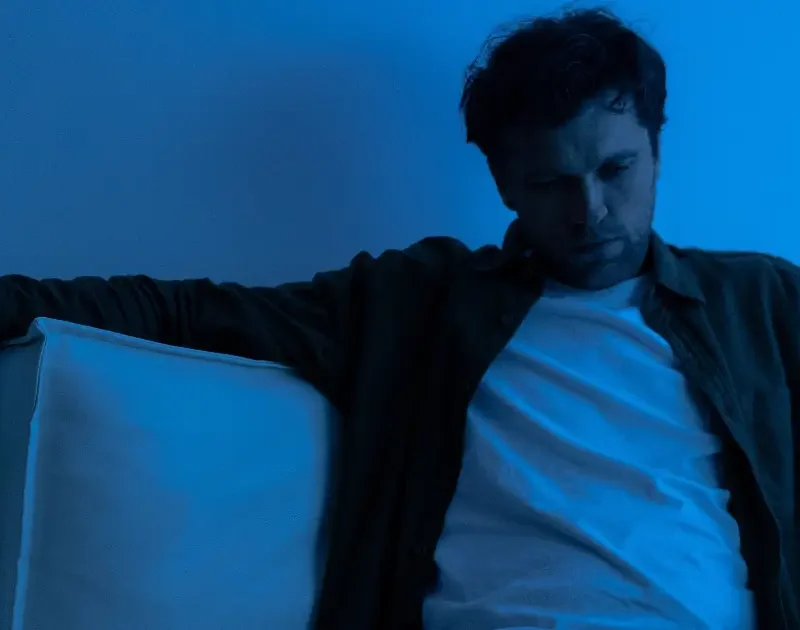 Nachdenklicher Mann sitzt allein auf dem Sofa in blauem Licht, Sinnbild für emotionale Rückfälle.
